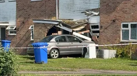 A car that has collided into the front of a house. A cordon has been placed around the crash. It is a terraced house and there are blue wheelie bins in the front gardens.