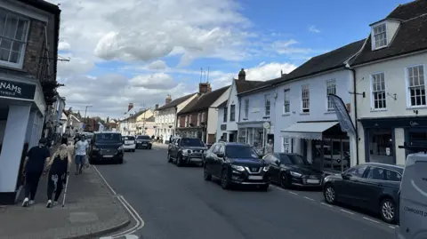 Ben Parker/BBC Hadleigh High Street with cars parked on both sides of the road, vehicles are travelling in both directions. People are walking on the pavement. Shops line the street.
