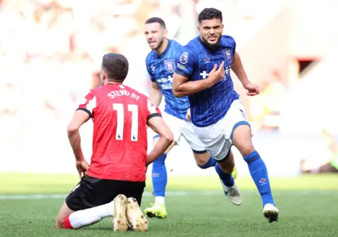 Getty Images Sam Morsy celebrates in his full Ipswich Town blue and white kit, with an enthralled facial expression, seen running past a Southampton player on his knees. He is frustrated, with his hands on his hips.