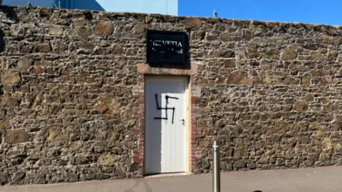 A wide shot of a a white door with a black spray-painted swastika over it.