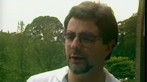 A screengrab of BBC Archive footage of Gibney. He has thick brown hair, thin rimmed glasses and a beard. He wears a white shirt.