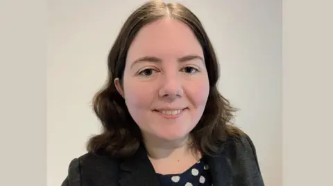 University of Northampton Alice Wilby with wavy shoulder length brown hair looks directly at the camera. She is standing inside.
