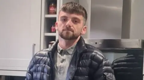 Jordan Parlour, a 28-year-old man with short brown hair and beard pictured in front of kitchen cupboards