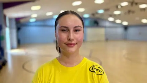 Ellie Drennan-Shoti pictured with slicked back dark hair and she's wearing a yellow t-shirt. Behind her is the roller skating rink which is mainly a wooden floor with painted lines on it.