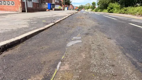 A road where the surface has been damaged. There is a pavement either side as well as houses one the left hand side and trees on the right hand side.