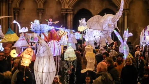Project Factory A procession of people holding up home-made lanterns in different animal shapes that are lit up.