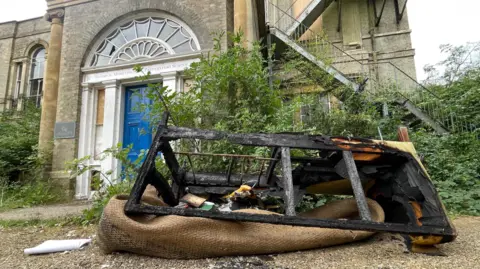 A burnt out sofa frame in front of Moreton Hall School. The blackened frame is resting on a piled up brown possible rug and there is other burnt debris resting on the rug. Behind the debris is the portico-style front door of the Georgian-style manor house. There are a lot of weeds and overgrown plants on either side of the front door. 
