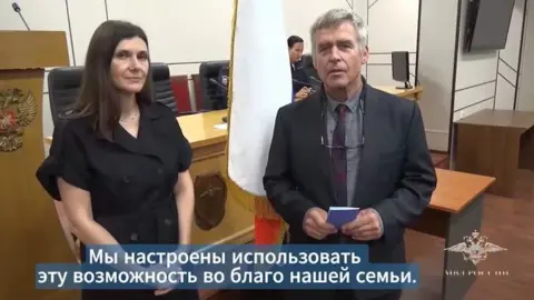Russian Ministry of Internal Affairs Two people in formal clothing stand at the front of a courtroom, one holding a small blue booklet and folded glasses. A light-coloured flag on a pole stands between them. Wooden desks, chairs, and a lectern with an emblem are visible behind. A person sits at a desk in the background. A crest appears on the right side of the image.