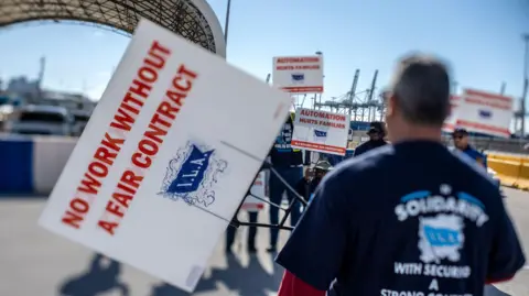 EPA-EFE Trabajadores portuarios asisten a una manifestación y sostienen carteles en el puerto de Miami, el primer día de la huelga de trabajadores portuarios de las instalaciones de carga de la Costa Este y del Golfo en Miami, Florida (EE.UU.), 01 de octubre de 2024. 