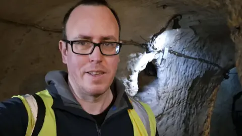 A view of a man with glasses and a hi vis vest stood in a grey cave.