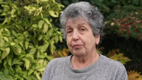 A woman with grey hair wearing a grey jumper is standing outside. Bushes are behind her filling most of the background.