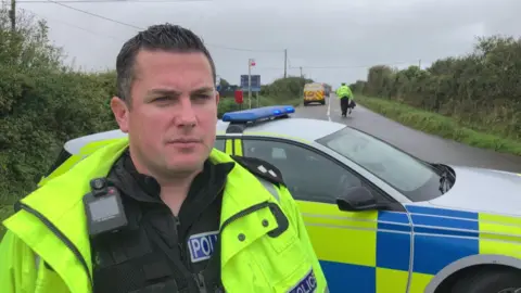 Tony Hannaford stood on the right wearing a yellow hi-vis jacket with a black jacket underneath. He is stood in front of a yellow and blue police car. In the background is a police officer walking on the right side of the road and a yellow and red police van on the left. There are hedges and signs either side of the road