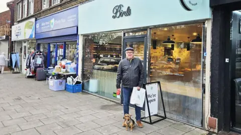 Supplied A man in a black jacket, blue flat cap, blue jeans and brown boots is stood in front of a bakery. He has a small dog on a lead in one hand and a carrier bag in the other. He is stood beside a swinging A-board sign which says 'We're open.' The bakery has a large glass front and a lime green sign which reads 'Birds.' The bakery is attached to a black barbers and a blue charity shop. There is another charity shop next to that further down the pavement.