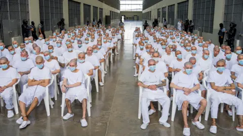 A group of hundreds of men with shaved heads, wearing white t-shirts and shorts or trousers and blue surgical masks, are seen sitting on white plastic chairs in an open space at a confinement centre in El Salvador