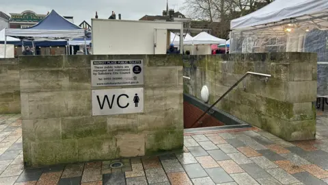 Market Square toilets in Salisbury. A staircase leads down to the block which is underground. There is a wall around the top of the block and a WC sign on the side. Market stalls can be seen in the distance.