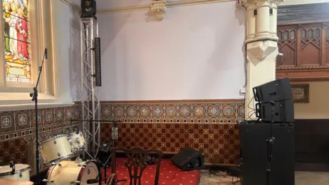 A stage area with red carpet and gold details. There is a black sound system and a cream coloured drum kit. On the back wall behind it is a colourful stained glass window
