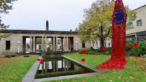 Stand Up For Southport Red knitted poppies hang down in a drape, with purple knitted poppies forming one giant poppy within the drape. It hangs down in Remembrance gardens in Southport, on an autumn day