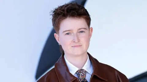 Getty Images A person with short reddish hair wearing a white shirt, colourful tie and a red leather jacket