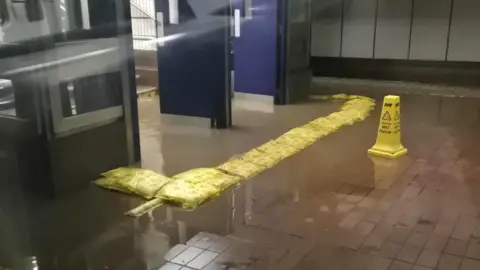 Flooding at the entrance to the train station. There are yellow sand bags lined up in front of the doorway and a yellow WET FLOOR cone.
