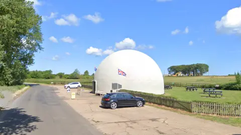 Google A white dome next to a road, with a Union Jack flag flying, cars parked and picnic tables on a grass area behind it