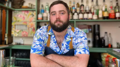 Man in front of a bar wearing a colourful blue shirt and apron