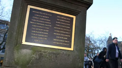 PA Media A stone plinth located outside in a tree-lined square, with a gold-rimmed black plaque that features 21 names in gold lettering. The general public can be seen on the right walking past the sign.