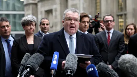 Reuters Tom Watson in dark suit speaking off notepad in front of a row of microphones. People in suits are standing behind him.