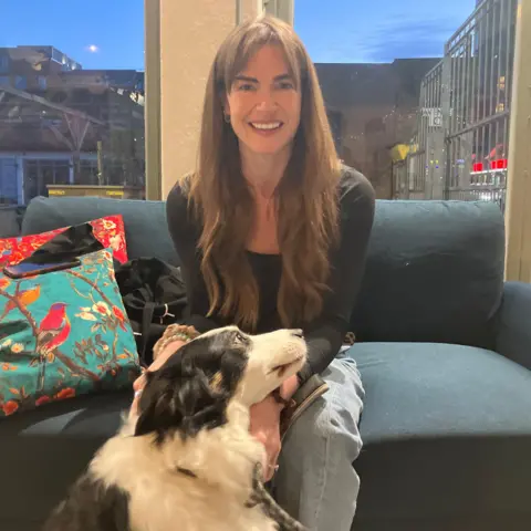 A woman with long brown hair sits on a blue sofa with a black and white border collie dog resting her head on her lap
