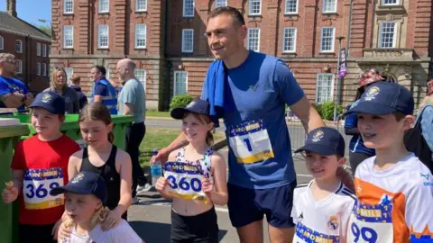 BBC/Cathy Killick Kevin Sinfield in the centre, wearing a blue t-shirt and blue shorts, with a blue towel on his right shoulder. He is posing with six young girls who are wearing dark blue caps and wearing sports clothing with number badges.