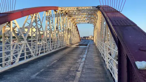 A view at one end of a semi-circular arched steel bridge with a tarmac road. The top beams are dark red and the intricate steelwork is an off-white. The sky is blue with no clouds.