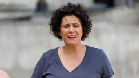 PA Media A woman with shoulder-length black hair. She is wearing a blue/grey top and appears to be standing outside. 