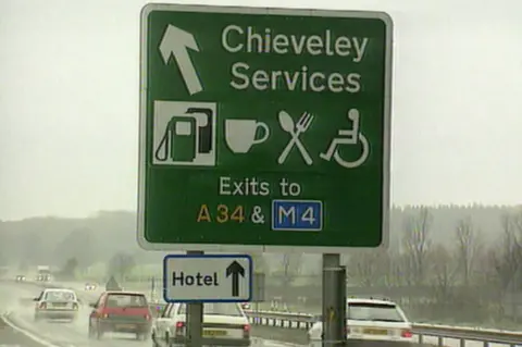 A green motorway Chieveley Services sign with white images and lettering on the carriageway where cars can also be seen. 