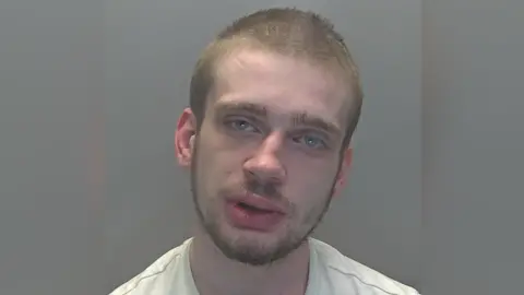 Custody image of Moorcroft against a grey background. His head is leaning slightly to the right and he has light brown facial hair and short hair. His eyes are blue and he is wearing a white top. 