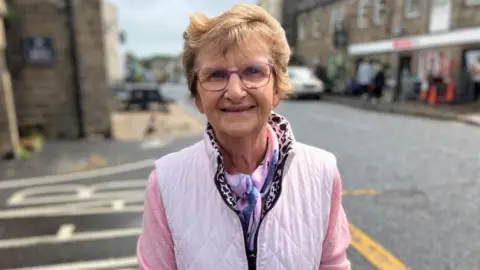 BBC/Seb Cheer A woman, Yvonne, smiles at the camera. She is wearing a pink top, lighter gilet, purple scarf and purple glasses. She stands in front of a market town street.