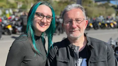 Stuart Woodward/BBC Holly McAllister is on the left. She has black hair with green highlighted strands. She wears a black quarter-zip top and has black-rimmed glasses on. Her father, Andrew, is on the right. He has short grey hair and a stubbly grey beard and silver-rimmed spectacles. He is wearing a black jacket over a black T-shirt. Both are smiling at the camera. There is a row of motorbikes in the background.