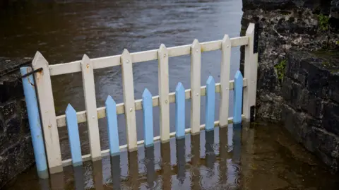 Ben Stevens/Getty Images A blue and white gate closed gate is seen with flood water half way up the gate which is attached to a stone wall on either side.