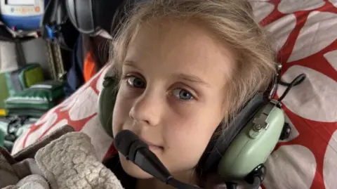 A young girl is looking at the camera. She is wearing a green headset which has a microphone attached. She is in a helicopter and is resting on a pillow while surrounded by medical equipment.