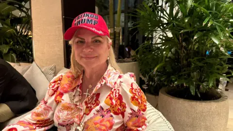 Maria McNally is photographed wearing a red hat, floral blouse and a large gold cross supporting Trump 