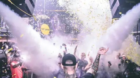 The picture shows a packed nightclub scene during a high‑energy moment in the middle of a DJ set. The photo is taken from behind the DJ booth, with the DJ in the foreground facing the crowd. Smoke cannons fire bursts of white fog across the room, and yellow confetti fills the air. People in the crowd are holding up their phones, filming the atmosphere and the performance. Lights, speakers and balconies full of people frame the scene, and the venue logo “THE DEPØ” appears in the bottom right corner.