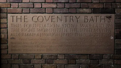 Aaron Law A sign on a brown brick wall which reads The Coventry Baths and has more information written below