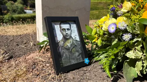 A framed picture of a war soldier is standing up on a grave. Behind is a headstone, and flowers.
