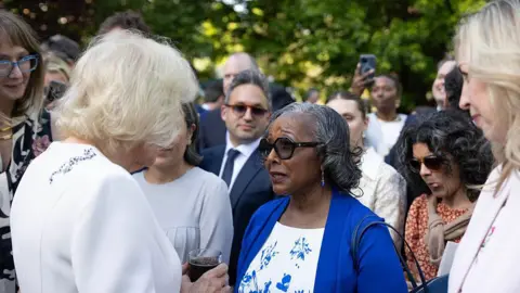 Getty Images  Queen Camilla in a white dress holds a glass as she speaks to president and CEO of domestic violence charity House of Ruth Sandra Jackson in a white dress and blue jacket at the British Embassy garden party in Washington. Other guests are in the background. 