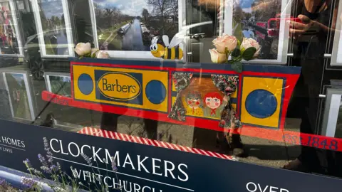 Whitchurch Waterway Trust A miniature wooden narrowboat painted in bright colours displayed in the front window of a shop, with drawings of the cartoon characters Rosie and Jim inside.