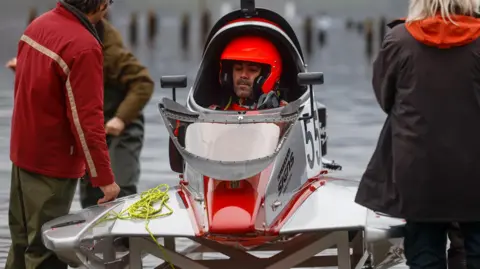 A man in a red helmet is pictured sitting inside a white and red powerboat near the water's edge. The boat has the number 55 on it. There is a blonde-haired woman to the right of the boat and two men to the left. The man in the boat is looking nervous. The boat is curved and has wings. 