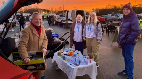 Phillip Norton/BBC Look North presenter Peter Levy pictured during his mug giveaway in Hull. He is wearing a long brown coat and is taking a box of mugs out of his car. He is smiling at the camera. There are several people standing at the stall.