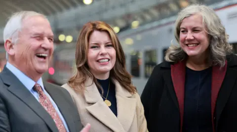 Iain Buist/NCJ Media Glen Sanderson and Kim McGuinness smile next to transport secretary Heidi Alexander. Sanderson has short white hair and is wearing a grey suit, pale blue shirt and red and mustard-coloured tie. McGuinness's red hair reaches just below her shoulders. She is wearing a black top, beige coat and a round gold pendant. Alexander's grey hair curls at the ends. She is wearing a black top and coat and a red scarf.