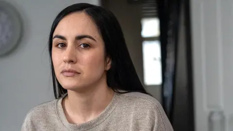 BBC Leanne Lucas, a woman with long, dark hair and dark eyes, wearing a grey top. She is looking at the camera as she sits in a living room with a door ajar behind her, with a view out to the hallway and the front door.