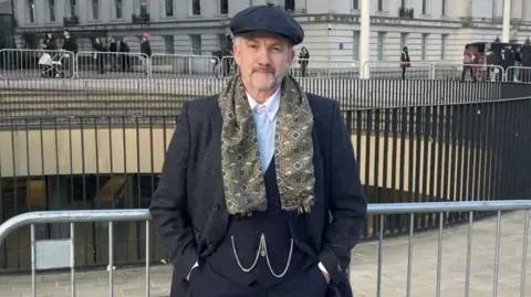 Arthur Cocker, a man dressed in a dark-coloured waistcoast and jacket with a white shirt and a dark-coloured flat cap with a patterned scarf. He is standing by railings in a city centre.