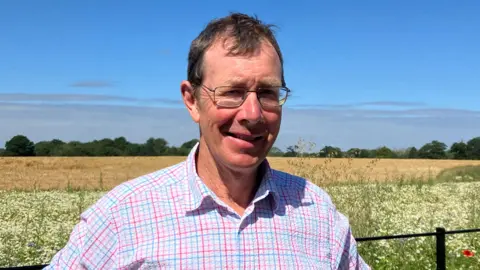 GUY CAMPBELL/BBC Farmer wearing a shirt and glasses stands in front of a field