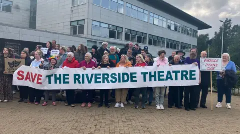 A number of people are seen standing outside the Causeway Coast And Glens Borough Council offices in Coleraine. They are dressed in colourful coats and jackets. A group is holding a banner that reads: 'Save The Riverside Theatre' and one man is holding a sign that reads 'Riverside Theatre Club'.